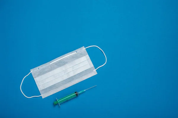 Green syringe and surgical mask with rubber ear straps. Typical 3-ply surgical mask to cover the mouth and nose. Procedure mask from bacteria. Protection concept. On blue background.