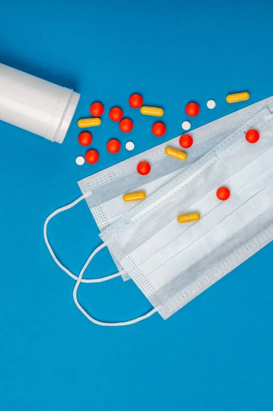 Red and yellow pills from white box and surgical mask with rubber ear straps. Typical 3-ply surgical mask to cover the mouth and nose. Procedure mask from bacteria. Protection concept.