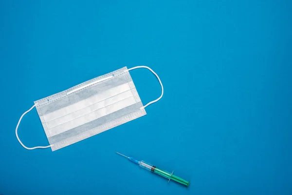 Green syringe and surgical mask with rubber ear straps. Typical 3-ply surgical mask to cover the mouth and nose. Procedure mask from bacteria. Protection concept. On blue background.