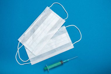 Yeşil şırınga ve lastik kulaklıklı iki cerrahi maske. Ağız ve burnu örtmek için tipik 3 katlı cerrahi maske. Bakterilerin prosedür maskesi. Koruma konsepti. Mavi arkaplanda.