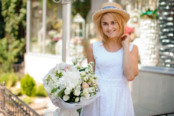 Chica en un vestido blanco y un sombrero de paja sostiene ramo festivo — Foto de Stock