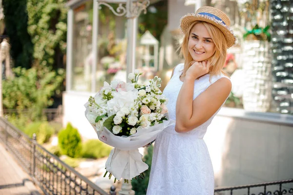 Chica en un vestido blanco y un sombrero de paja sostiene un ramo festivo — Foto de Stock