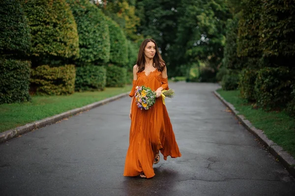 Atractiva mujer en vestido naranja caminando por la carretera con ramo de flores — Foto de Stock