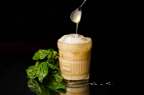 Bebida alcoólica fresca com espuma e gelo em um copo servido na superfície escura com manjericão — Fotografia de Stock