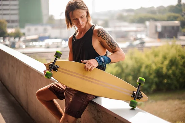 Atraente cara de cabelos longos sentado no parapeito com longboard — Fotografia de Stock