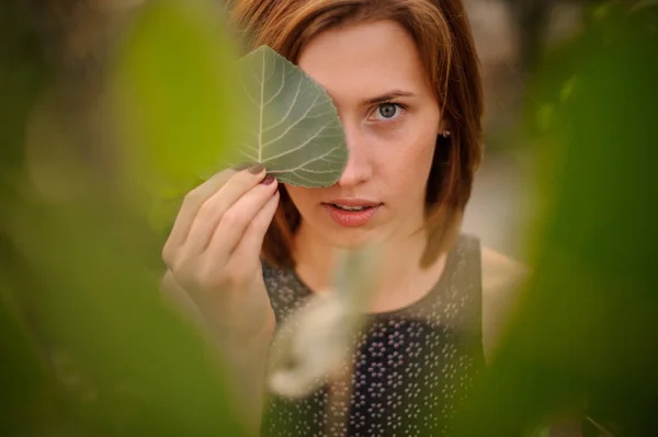 Привлекательная рыжая девушка, скрывающая половину своего лица зеленым листом — стоковое фото