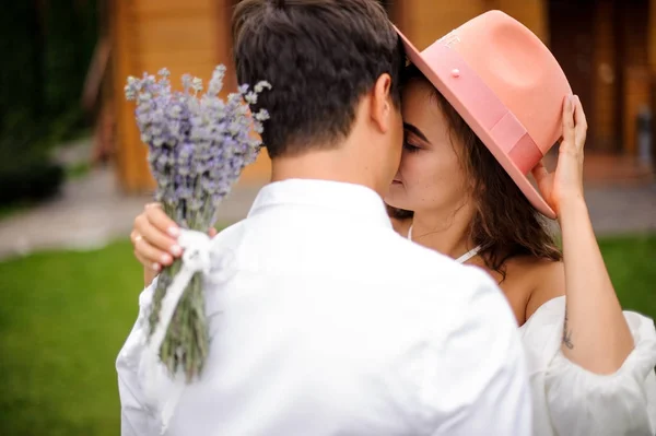 Sposo in camicia bianca che abbraccia sposa in abito bianco e cappello rosa con mazzo di fiori — Foto Stock