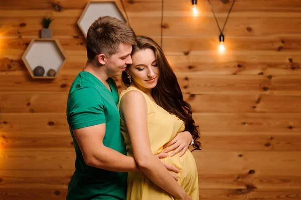 Happy man hugging his beautiful pregnant wife on the background of garland — Stock Photo, Image