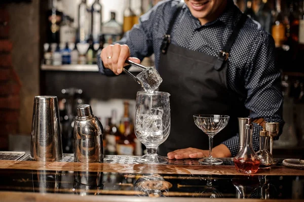 Barman colocando um grande cubo de gelo no copo com a ajuda de pinças de gelo especiais — Fotografia de Stock
