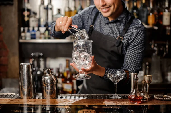 Barman sorridente colocando um grande cubo de gelo com a ajuda de pinças de gelo especiais — Fotografia de Stock