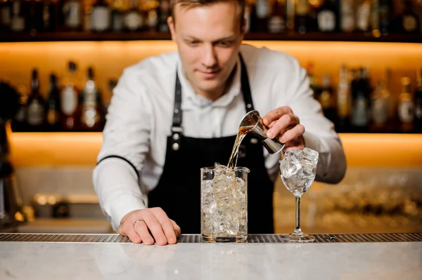 Joven barman vestido con una camisa blanca vertiendo bebida alcohólica en un vaso con cubitos de hielo — Foto de Stock