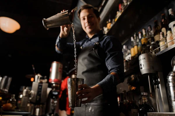 Jovem barman sorrindo derramando bebida alcoólica de um copo de metal em outro — Fotografia de Stock