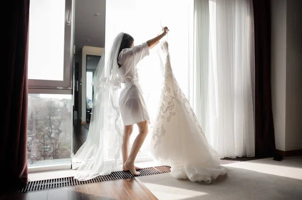 Nevěsta v županu a závoj drží svatební šaty na závěs — Stock fotografie