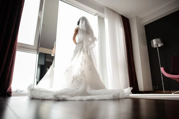 Sposa in abito da sposa e velo di seta guardando attraverso la finestra — Foto Stock