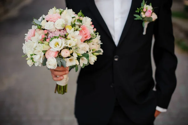 Noivo em um terno preto com boutonniere segurando um buquê de casamento — Fotografia de Stock