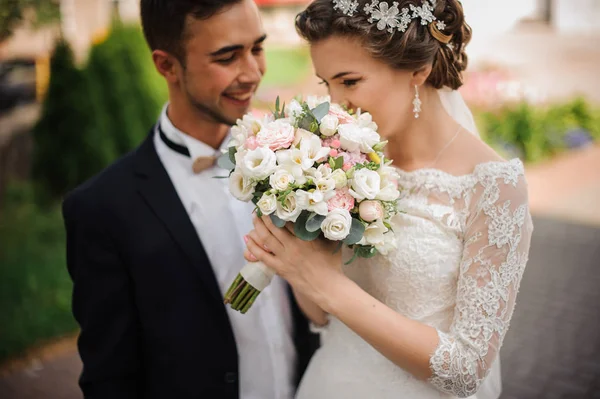 Braut genießt den Duft eines Brautstraußes, der Bräutigam steht lächelnd daneben — Stockfoto