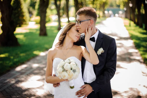 Noivo com óculos e noiva com um buquê de peônias gentilmente abraçar no parque — Fotografia de Stock
