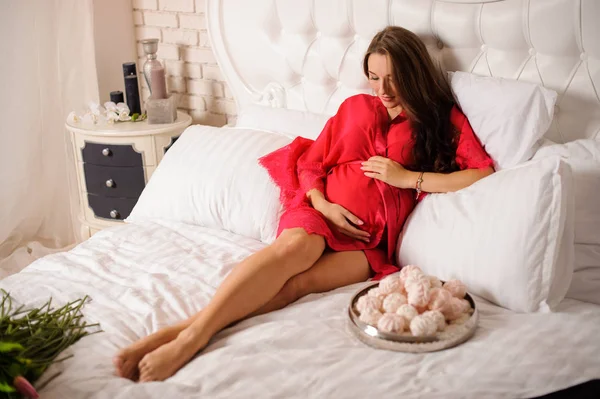 Piękna kobieta w ciąży ubrany w elegancki szlafrok leżącego na białym złe słodyczy — Zdjęcie stockowe