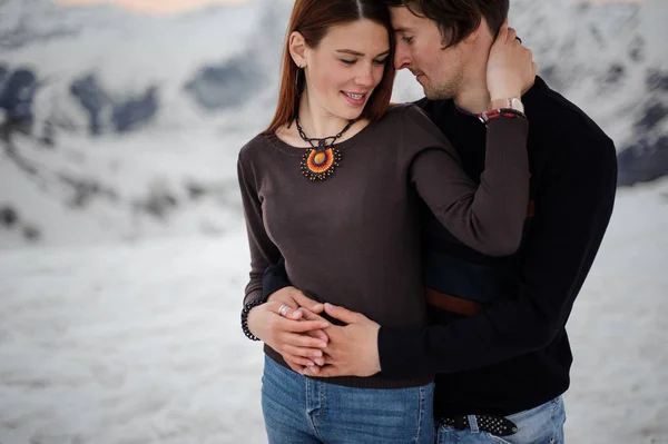 Jong koppel, in jeans en gebreide truien, knuffelen elkaar op de achtergrond van bergen sneeuw — Stockfoto