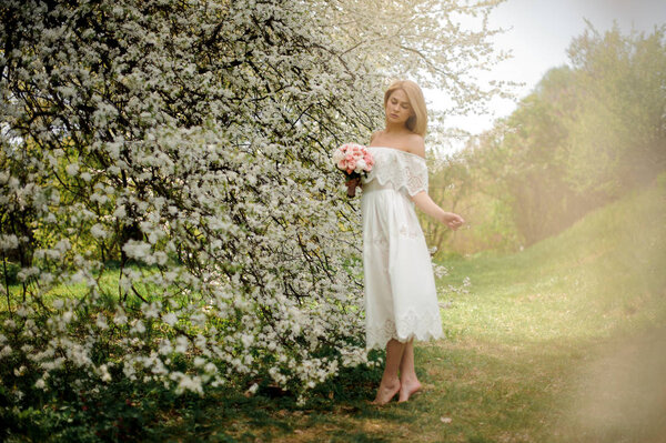 Beautiful blonde girl, dressed in a white dress, standing between branches of white blossom tree with a bouquet barefoot