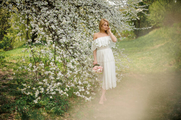 Beautiful blonde girl, dressed in a white dress with a open shoulders, standing between branches of white blossom tree with a bouquet