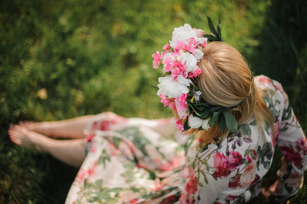Young and beautiful blonde girl in a flower diadem, dressed in a dress with roses , sitting on the grass