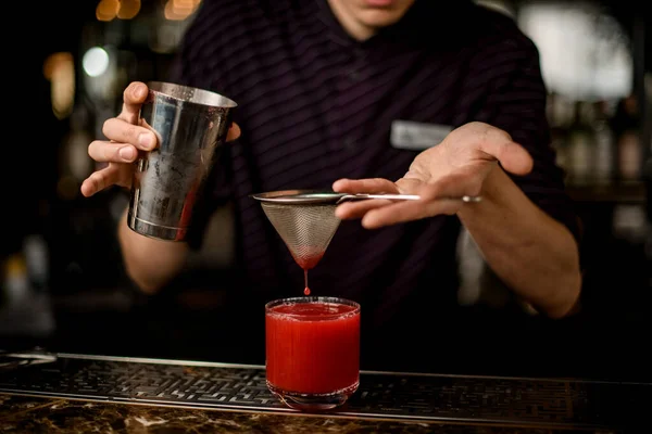 Cantinero profesional que vierte una bebida alcohólica roja del agitador de acero al vaso con hielo a través del tamiz — Foto de Stock