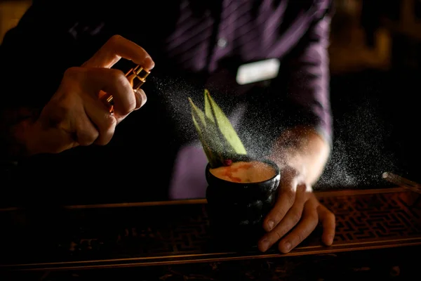 Barman spaying na černé sklenici s koktejlem zdobené tropickým listem, sušené růžové pupeny a esence kapky — Stock fotografie