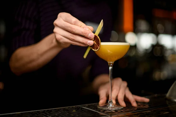 Professional bartender decorating yellow alcoholic cocktail in the glass with a dried lemon and tropical leaf