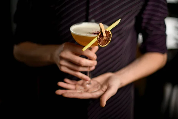 Bartender i lila skjorta som serverar gul alkoholhaltig cocktail i glaset med en torkad citron och blad i klädnypan — Stockfoto