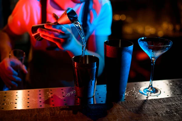Μπάρμαν ρίχνει ένα αλκοολούχο ποτό από το jigger σε ένα ατσάλινο σέικερ στο μπλε και κόκκινο φως — Φωτογραφία Αρχείου