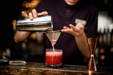 Profesyonel erkek barmen, kırmızı alkollü bir içeceği çelikten bardağa kadar süzgeçten buzla dolduruyor.