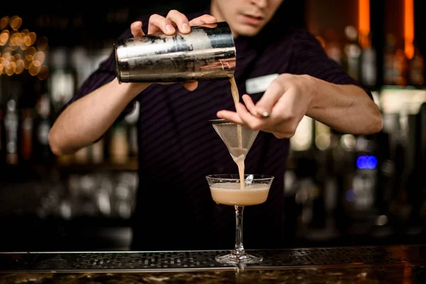 Männlicher Barkeeper, der ein alkoholisches Getränk aus dem Stahlshaker in das Cocktailglas durch das Sieb gießt — Stockfoto