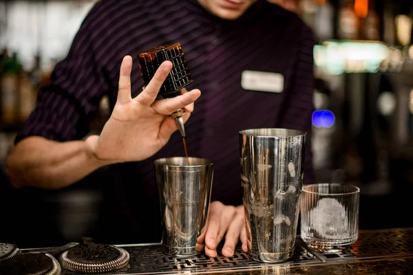 Professioneller Barkeeper, der einem alkoholischen Getränk in einem Stahlshaker einen braunen Bitter aus einer Glasflasche hinzufügt — Stockfoto