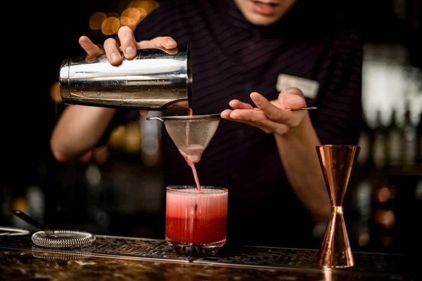 Barkeeper gießt ein rotes alkoholisches Getränk aus dem Stahlshaker in das Glas mit Eis durch das Sieb — Stockfoto