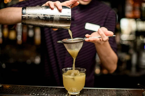 Männlicher Barkeeper, der ein gelbes alkoholisches Getränk aus dem Stahlshaker in das Glas mit Eis durch das Sieb gießt — Stockfoto