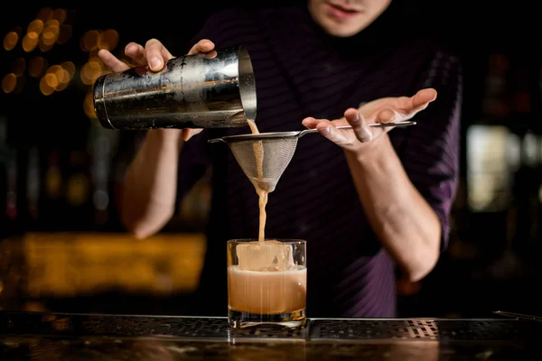Männlicher Barkeeper, der ein alkoholisches cremiges Farbgetränk aus dem Stahlshaker in das Glas mit Eiswürfel durch das Sieb gießt — Stockfoto