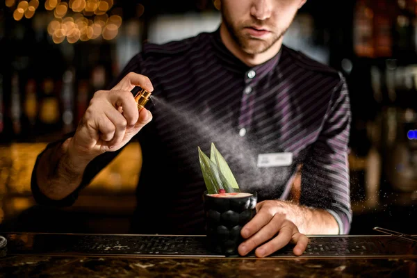 Professional bartender spaying on a black glass with cocktail decorated with a tropical leaf, dried rose bud and essence drops