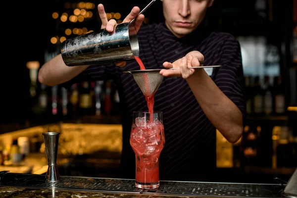 Professioneller Barkeeper, der ein rotes alkoholisches Getränk aus dem Stahlshaker in das Cocktailglas durch das Sieb gießt — Stockfoto