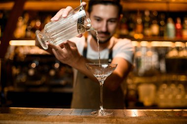 Barmen ölçü kabından bardağa süzgeç dökerek alkollü bir içecek dolduruyor.