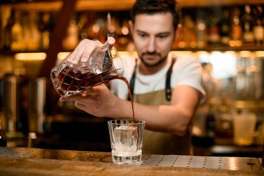 Profesyonel barmen, bardakla buzu karıştırıp kahverengi alkollü içeceği karıştırıyor.