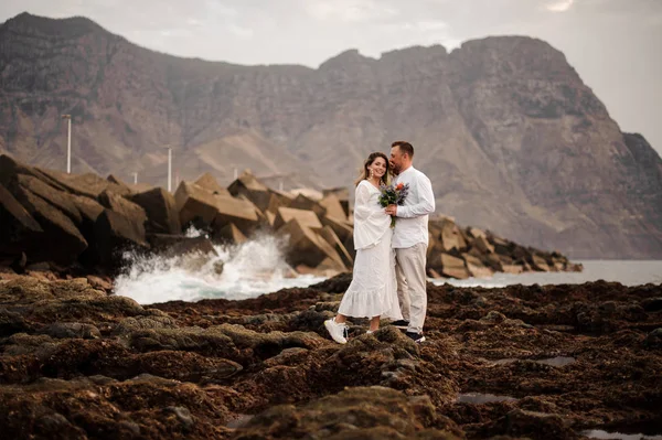 Muž ženich a žena nevěsta stojící na skalnaté pláži s kyticí — Stock fotografie