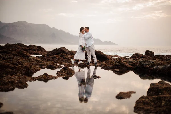 Muž ženich a žena nevěsta stojící na kamenné pláži s kyticí — Stock fotografie