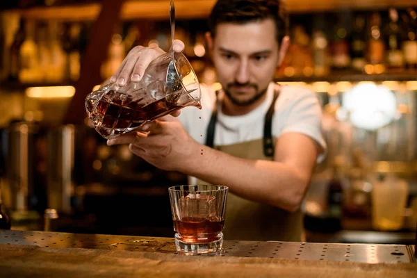 Barman masculino derramando uma bebida alcoólica marrom de vidro de mistura para um copo de coquetel com cubo de gelo — Fotografia de Stock