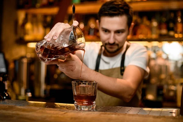 Бармен наливає коричневий алкогольний напій з суміші скла в коктейль з кубиком льоду. — стокове фото
