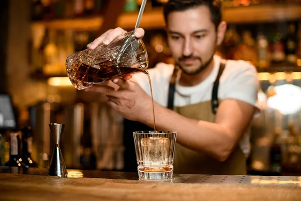 Cantinero vertiendo una bebida alcohólica marrón de la mezcla de vidrio a una copa de cóctel a través del tamiz —  Fotos de Stock
