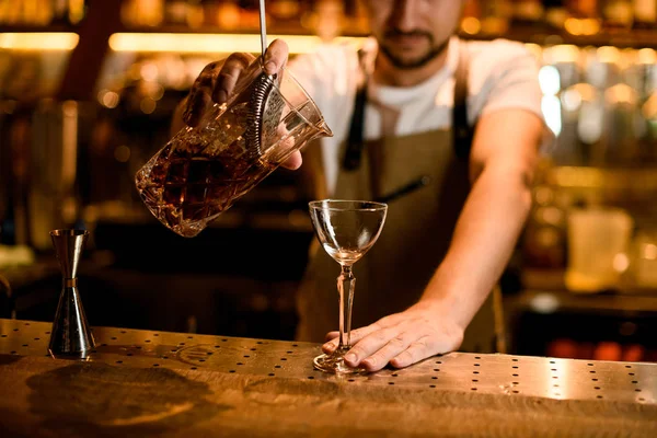 Чоловік бармен поливає коричневий алкогольний напій змішуючи склянку через сито — стокове фото