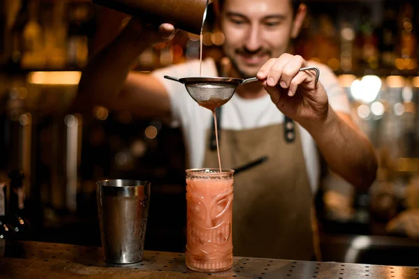 Usmívající se barman nalévá červený alkoholický nápoj z ocelového třepače přes síto — Stock fotografie