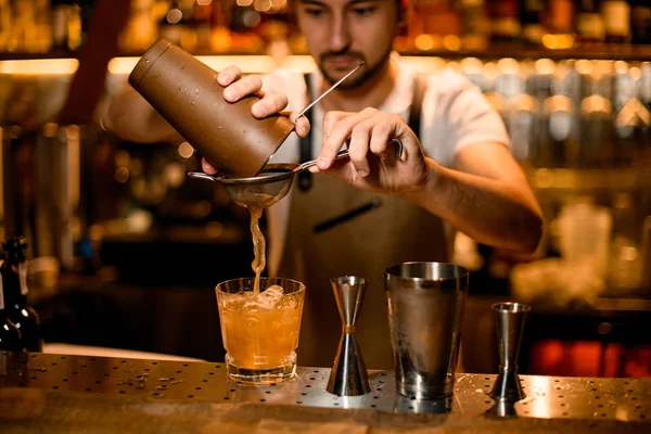 Професійний чоловічий бармен, що наливає апельсиновий алкогольний напій зі сталевого шейкера через сито — стокове фото