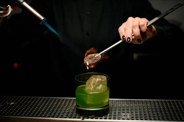 Барменша, подающая зеленый алкогольный коктейль в стакане добавляя кусочек лимона с твизерами сжигая его — стоковое фото